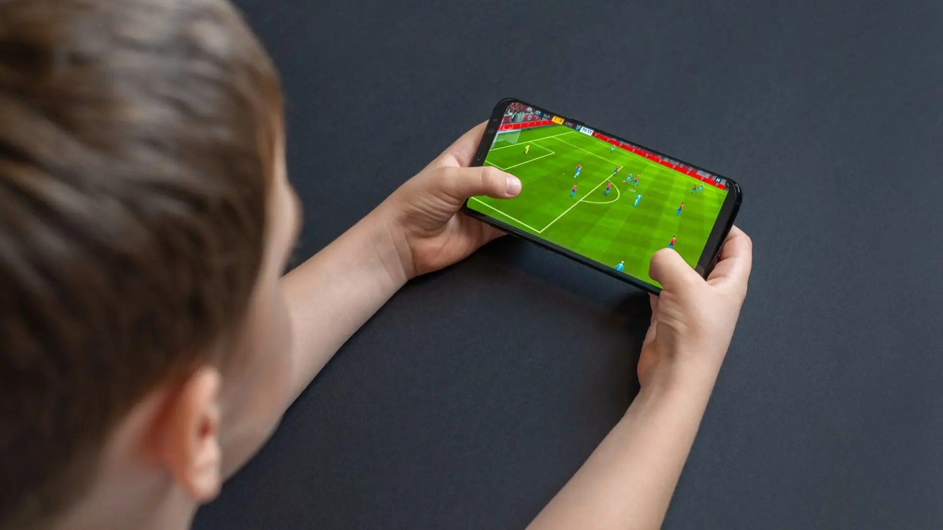 Criança com os dois braços sobre uma mesa segura um celular e joga Fifa Futebol 