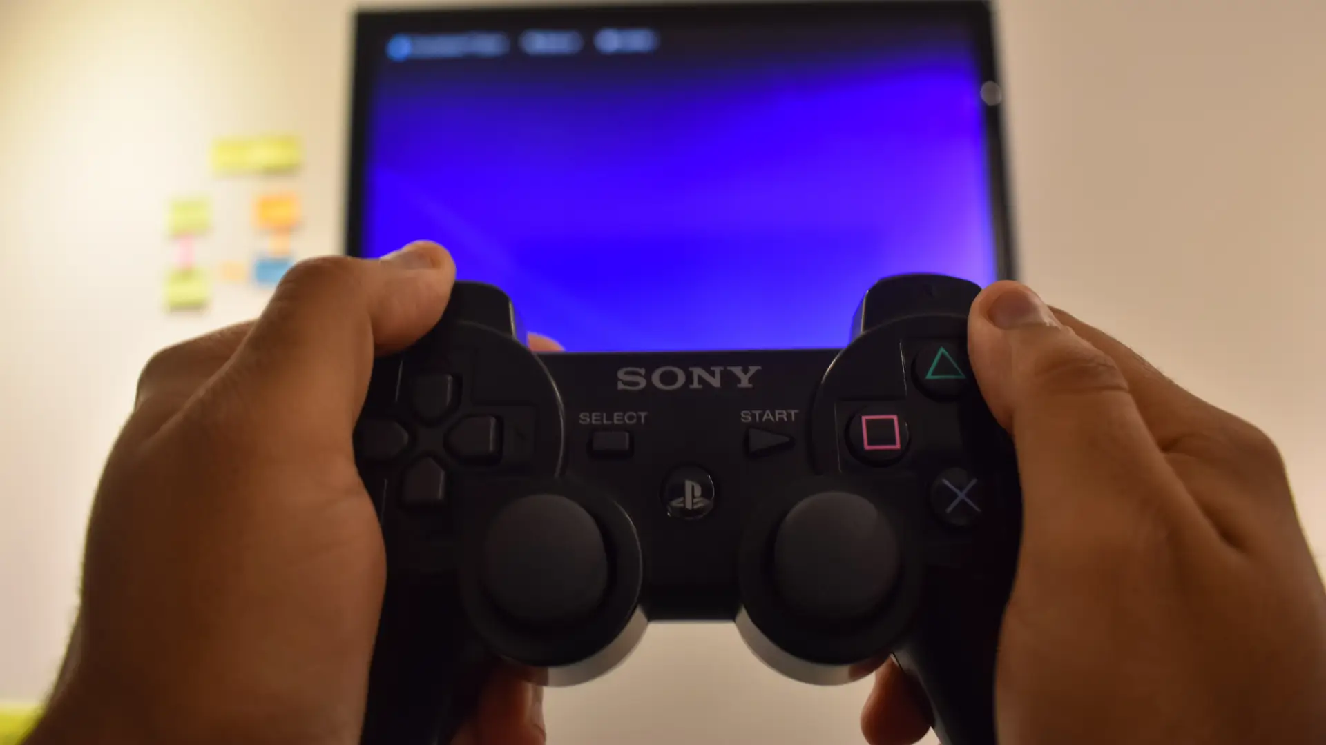 Mãos seguram um joystick de PS3, com a tela do console desfocada ao fundo.