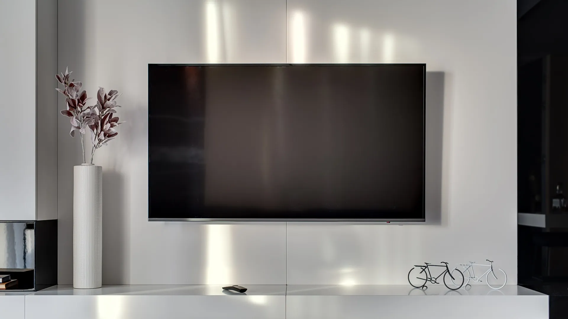 TV preta montada na parede de uma sala de estar, acima de um rack branco com decorações