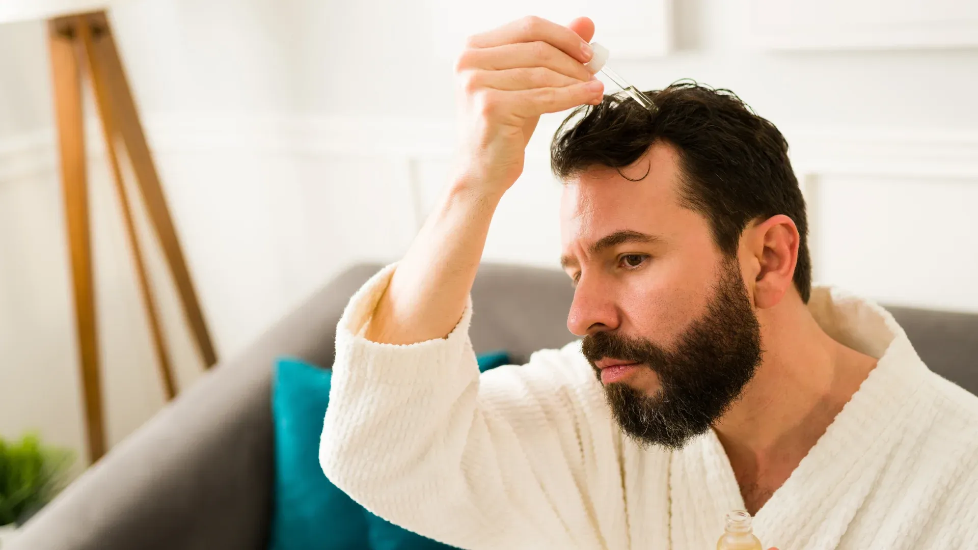 Homem branco com barba, vestido em um robe de banho, aplica um tônico na raiz dos cabelos.