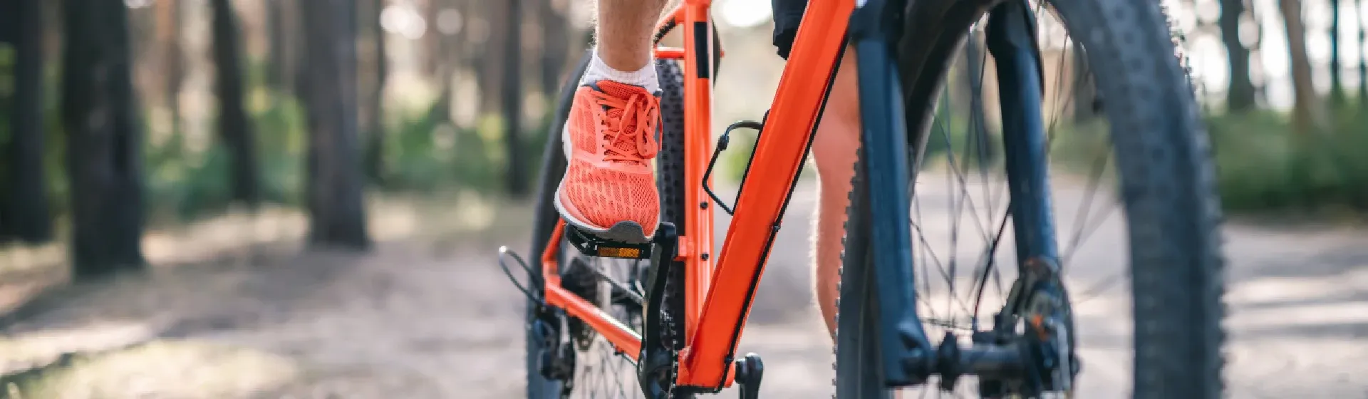 Foto das pernas de pessoa pedalando em cima de uma bicicleta laranja e com uma floresta no fundo