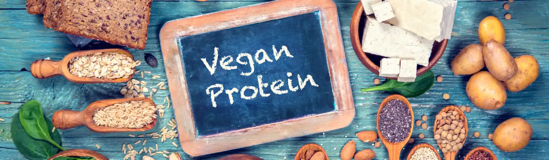 Em uma superfície plana está um quadro negro escrito “ vegan protein” com diversos vegetais e leguminosas ao redor dele.