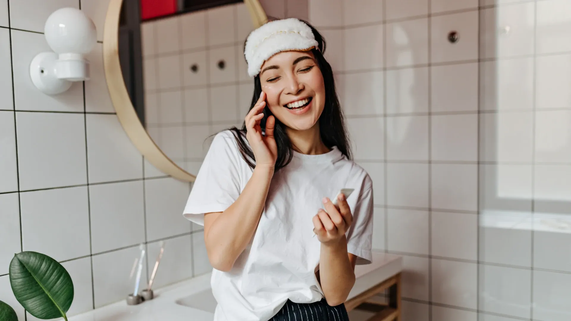 Mulher asiática sorrindo ao fazer skincare em banheiro branco