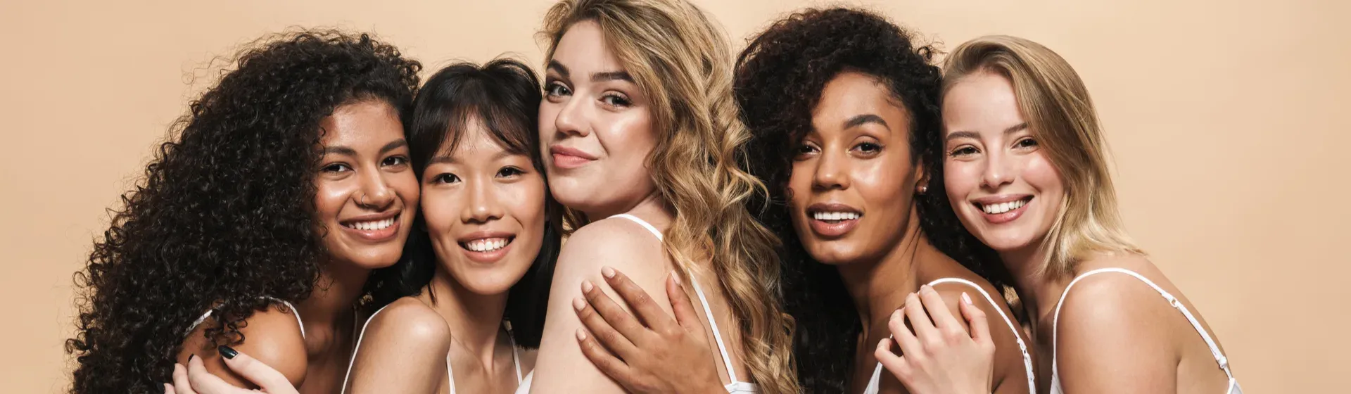 5 mulheres de cabelos e cores de pele diferentes, com camisas brancas, se abraçando e sorrindo em fundo bege 