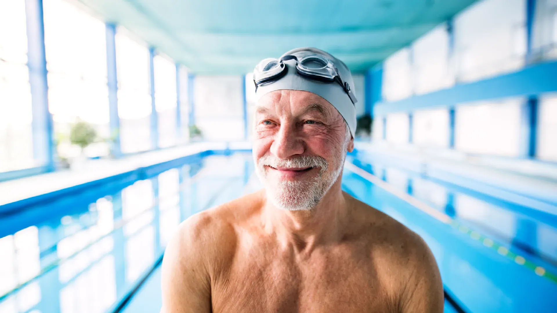 Homem idoso e branco com touca e óculos de natação sorrindo em frente à piscina