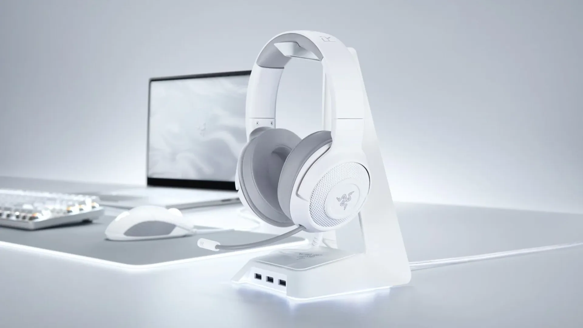 A imagem mostra um headset Razer Kraken branco, disposto sobre um suporte que está em cima de uma mesa com outros periféricos da marca
