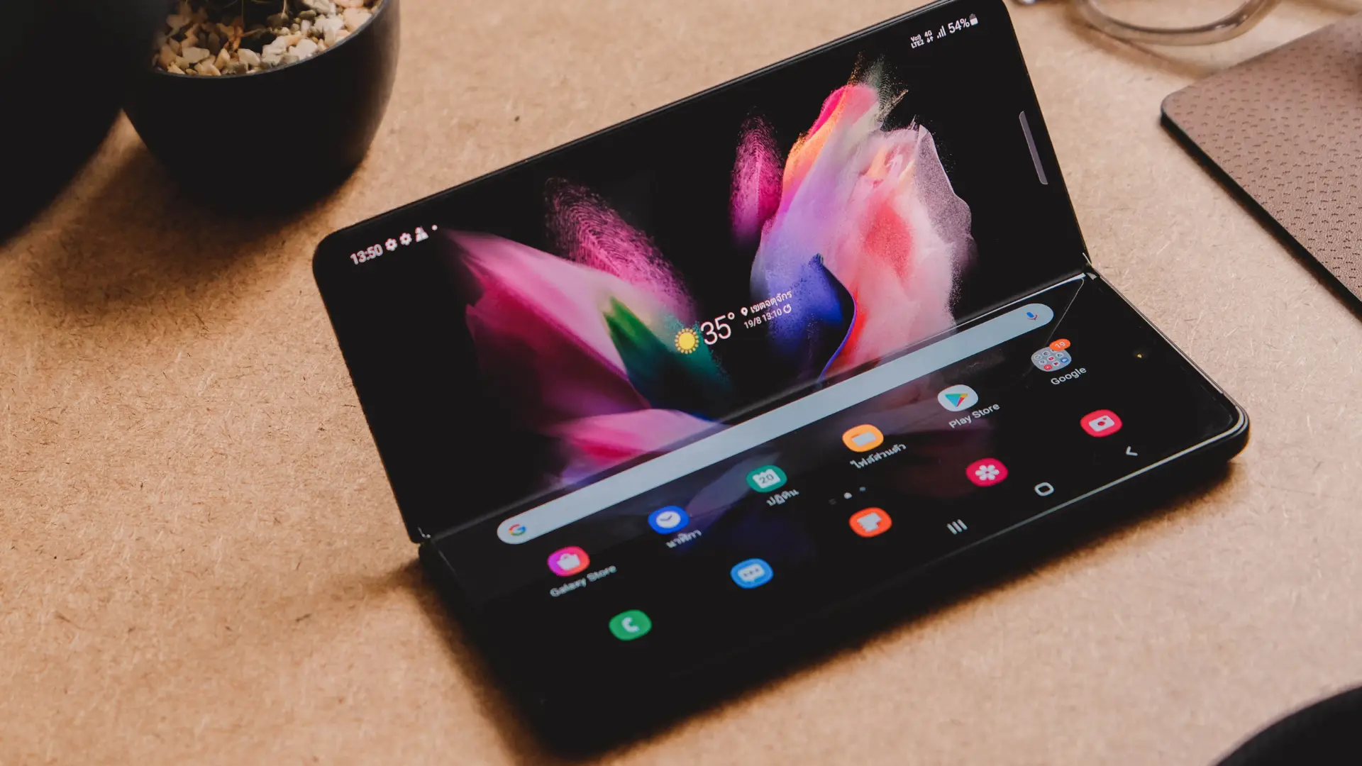 Um celular modelo Galaxy Z Fold 3 está com a tela aberta e ligada, sobre uma superfície, ao lado de diversos acessórios