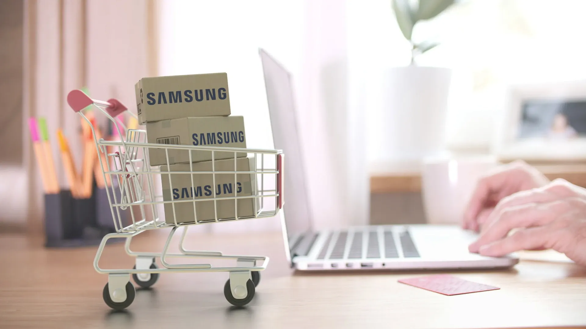 Imagem de um mini carrinho de compras com caixas escrito "Samsung" em primeiro plano, com uma pessoa usando um laptop no fundo