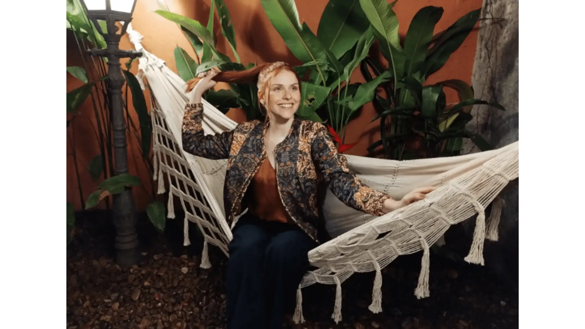 Uma mulher sentada numa rede com plantas ao fundo.