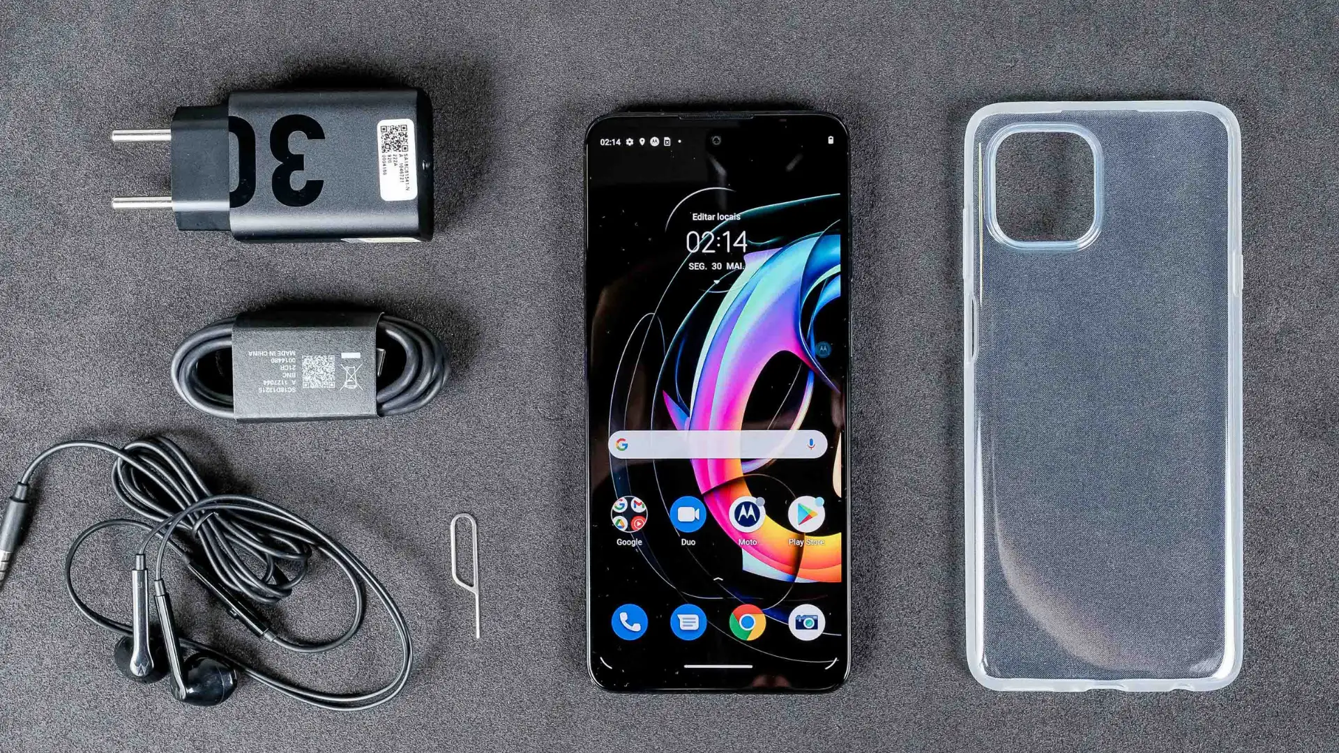 Motorola Edge 20 Lite no centro da foto com fundo cinza, capinha e carregadores em volta