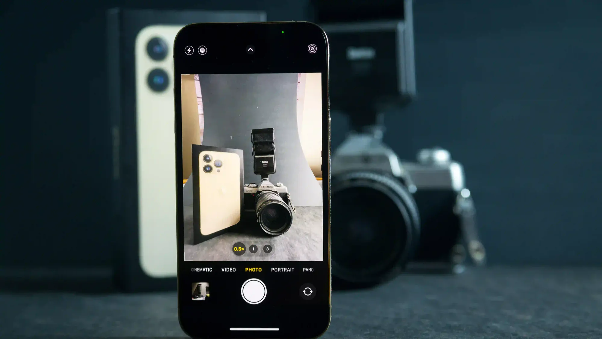 Um celular com a câmera ligada está em pé sobre uma mesa. Na tela aparece uma câmera fotográfica. Atrás do celular tem uma câmera fotográfica e ao lado um celular