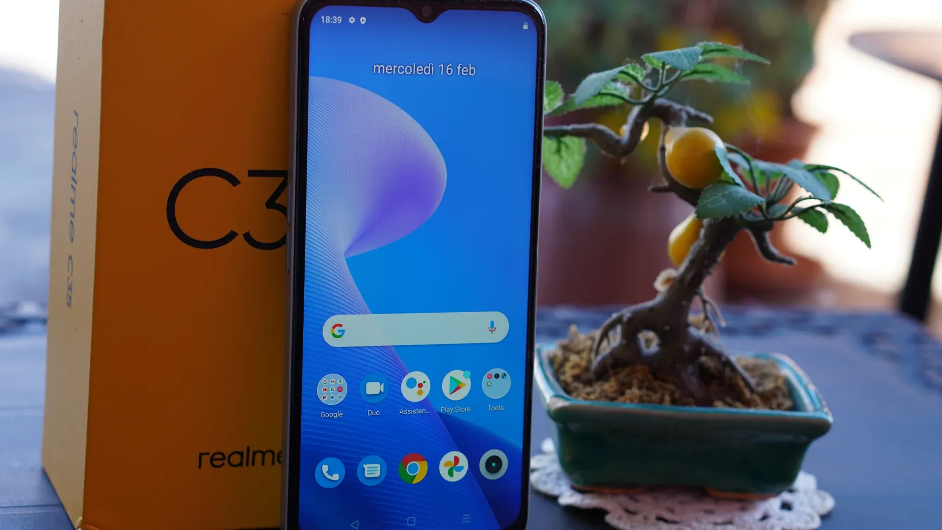 Um celular está em pé, em frente a uma caixa do mesmo aparelho e ao lado de um objeto de decoração