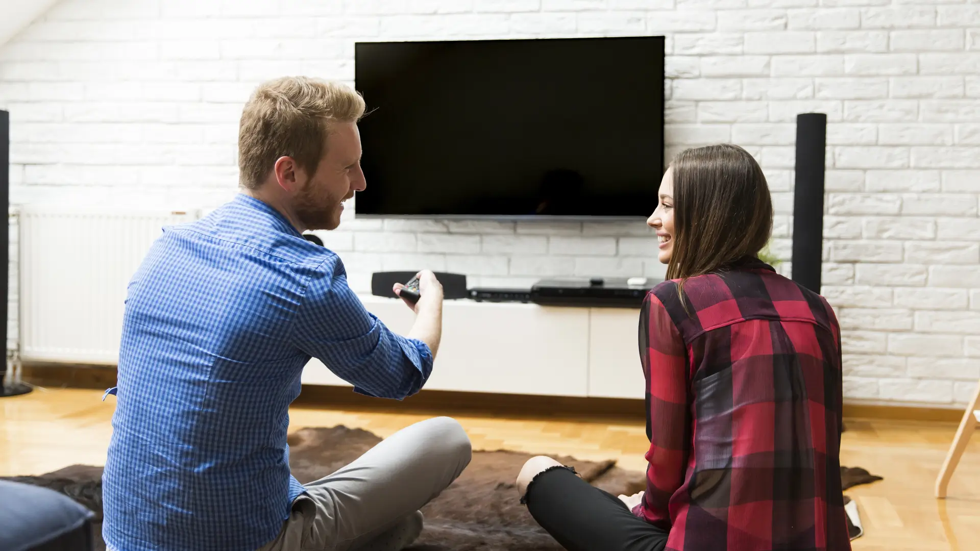 Casal sentado no chão da sala sorri enquanto aponta controle remoto para TV na parede