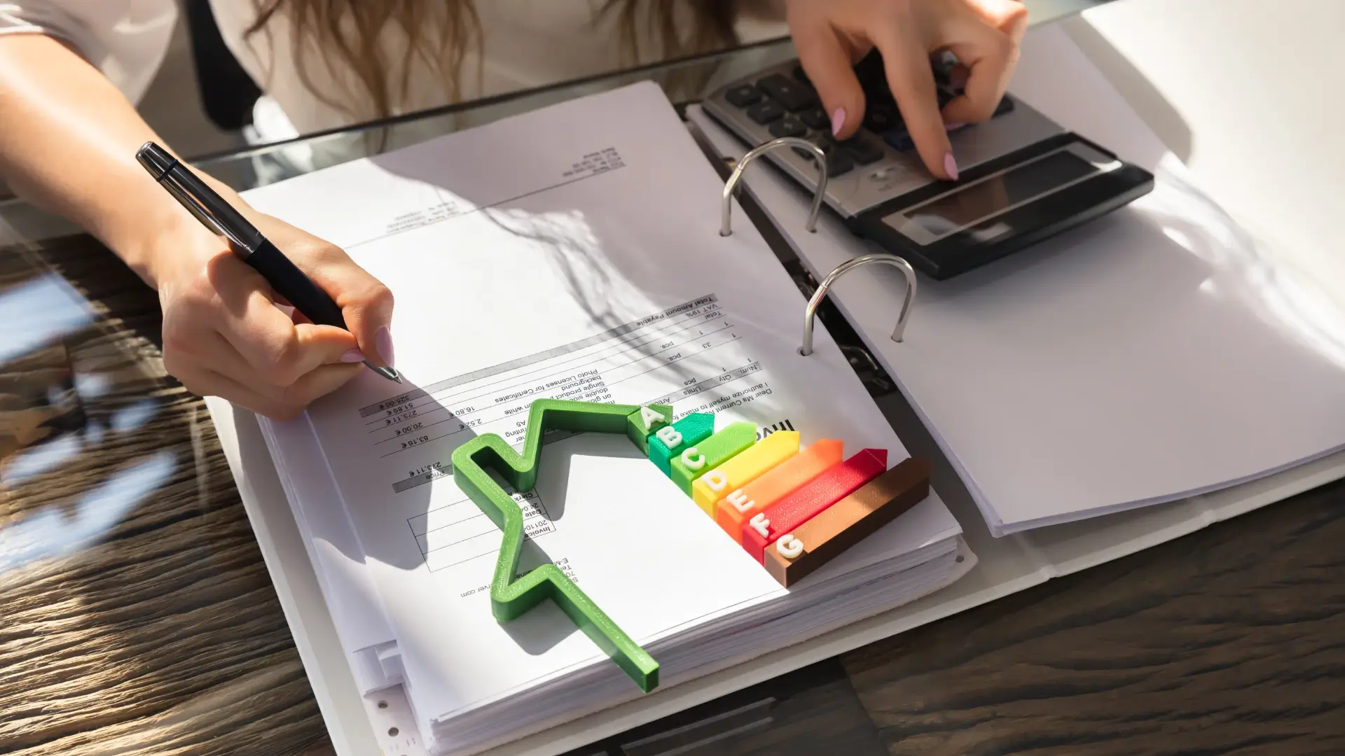 Close em mãos femininas fazendo contas na calculadora. Uma casinha com indicadores de selo procel A está sobre a mesa.