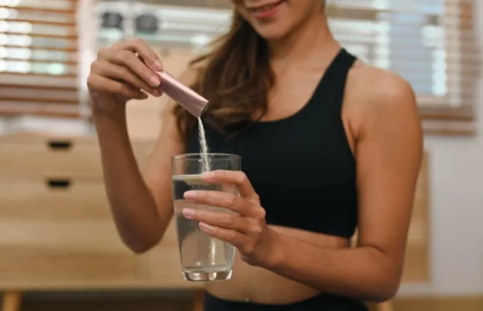 Mulher em roupa esportiva vertendo pó branco de um frasco rosa em um copo de água.