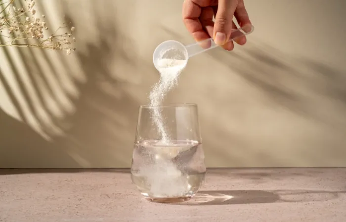 Mão despejando pó branco de uma colher de medição em um copo de água com luz natural.