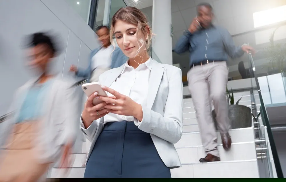 Mulher caucasiana mexendo em celular numa escada em ambiente corporativo