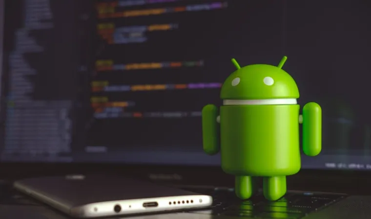 Celular deitado no teclado de um notebook com um boneco verde do Android ao lado.