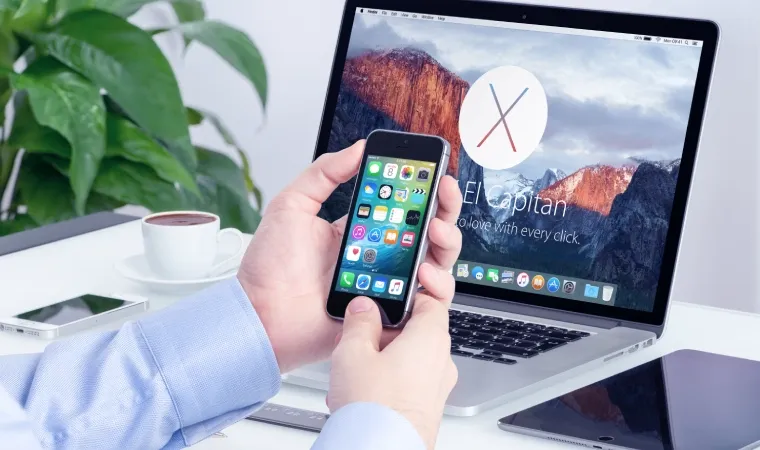 Pessoa mexendo em iPhone enquanto tem outro iPhone, uma xícara de café, um Macbook e um iPad em cima de uma mesa ao fundo