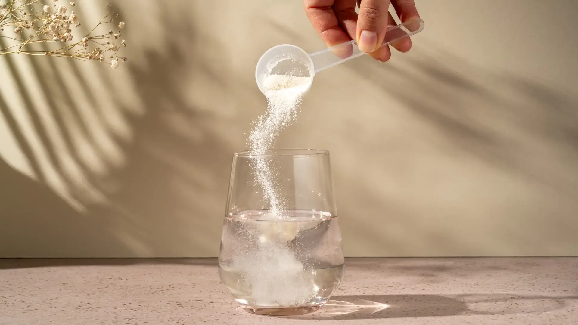 Mão feminina colocando um scoop de colágeno em copo de água apoiado na mesa