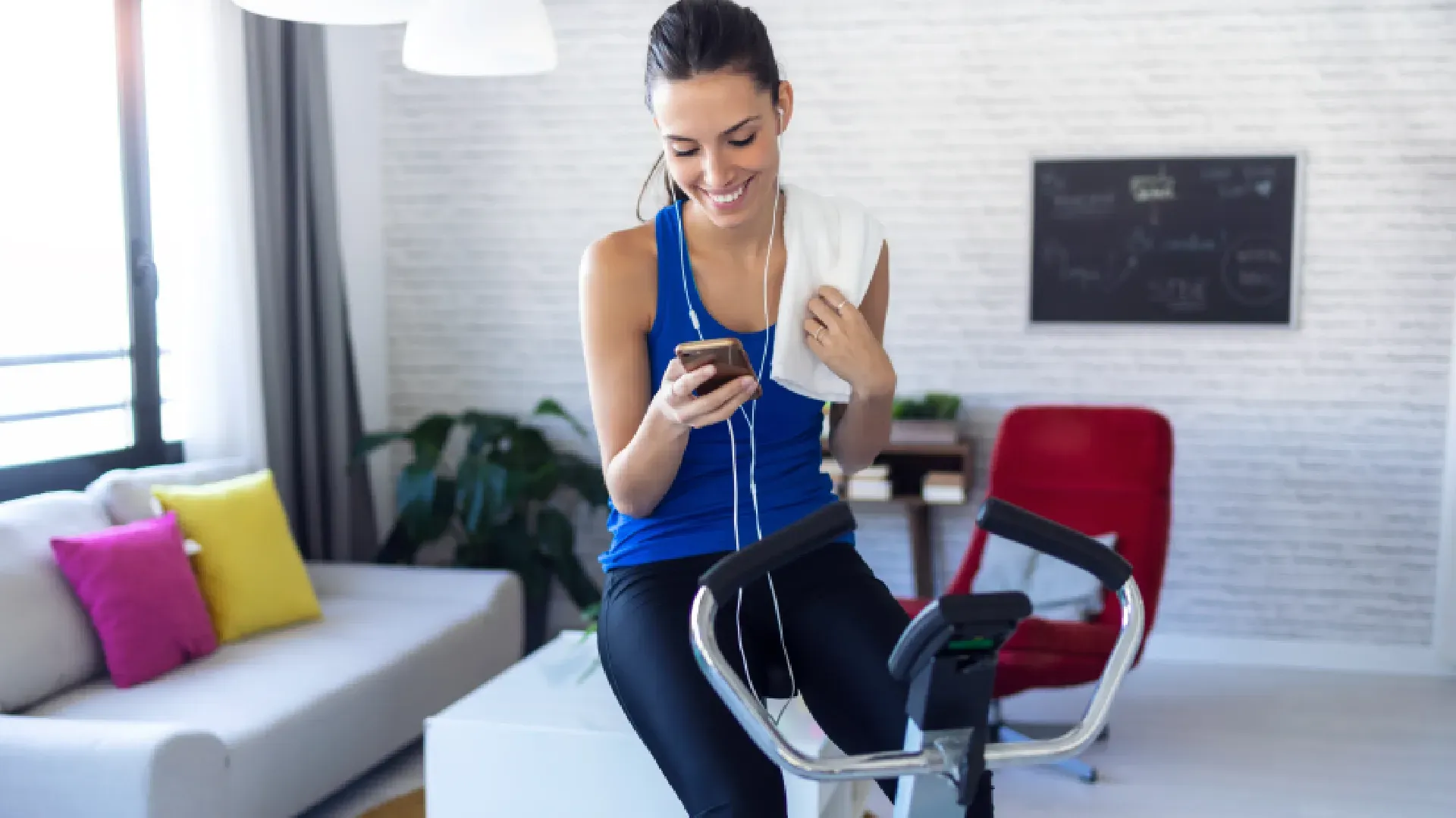 Mulher sorri mexendo no celular na sala de casa enquanto pedala em bicicleta ergométrica vertical