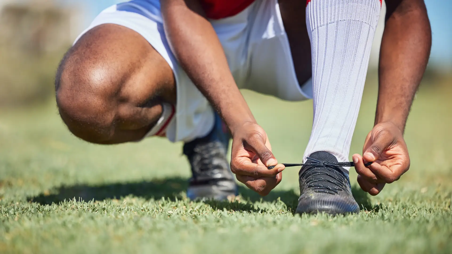 Close nas mãos de homem negro agachado em campo de futebol amarrando o cadarço de chuteiras pretas
