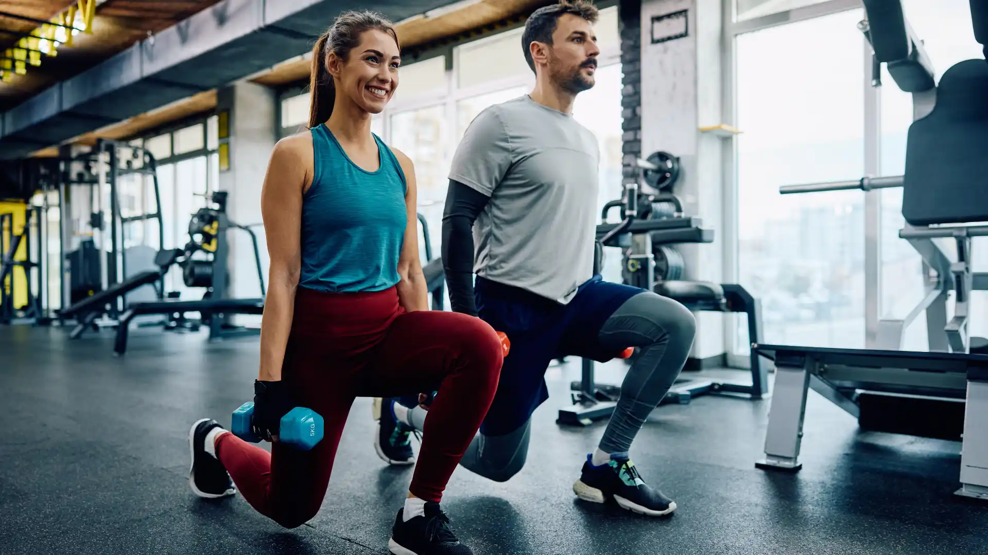 Um homem é uma mulher realizando um exercício com uma perna agachada segurando pesos com as duas mãos 