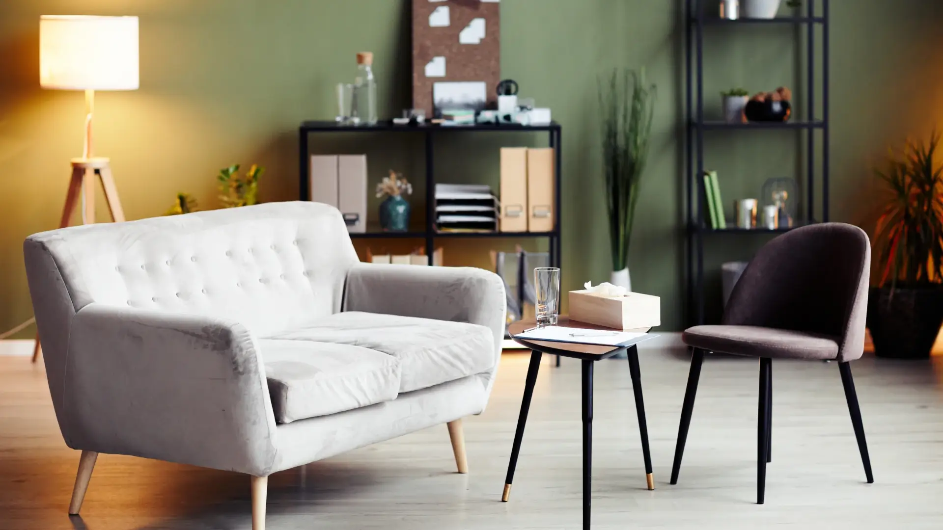 Sofá cinza de dois lugares, mesa e poltrona no centro do ambiente. Estantes ao fundo.