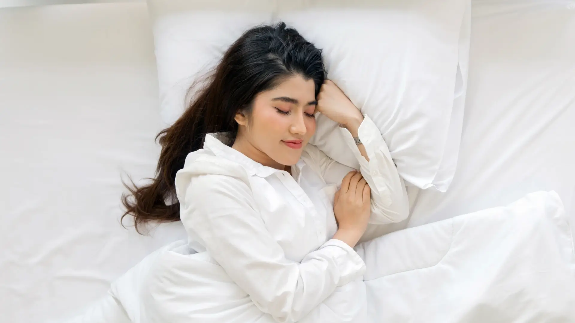 Mulher deitada na cama de olhos fechados, coberta por edredom branco. 