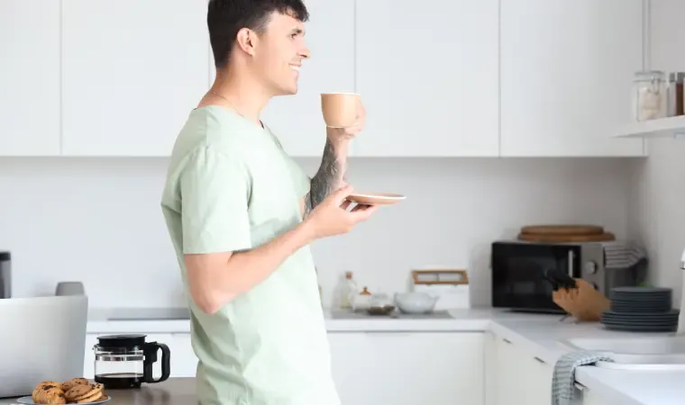 Homem após aprender como fazer café na cafeteira elétrica tomando café na xícara em pé na cozinha.