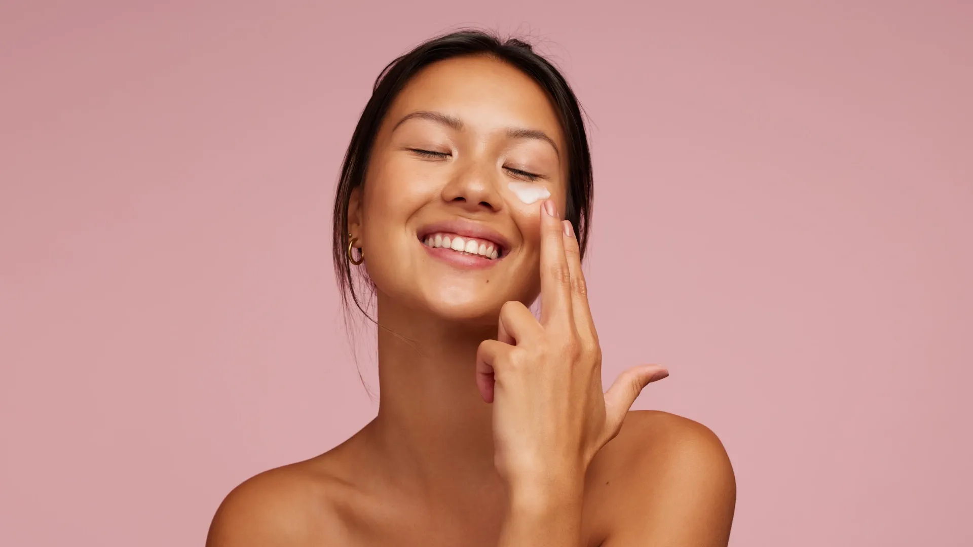 Mulher asiática sorrindo e aplicando produto na pele do rosto em um estúdio com fundo rosa