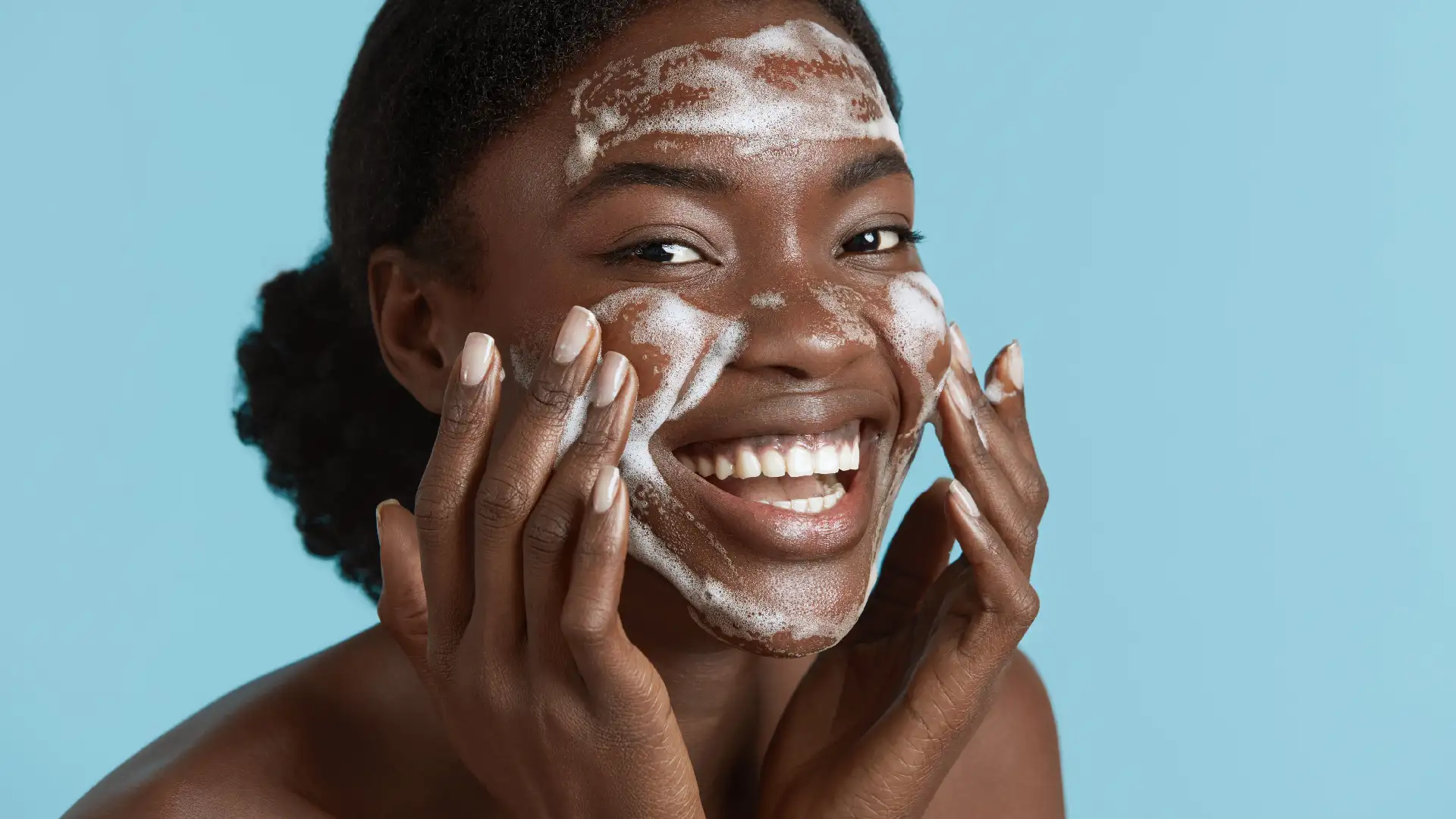 Mulher negra sorrindo enquanto espalha produto de limpeza facial branco e espumoso com as mãos