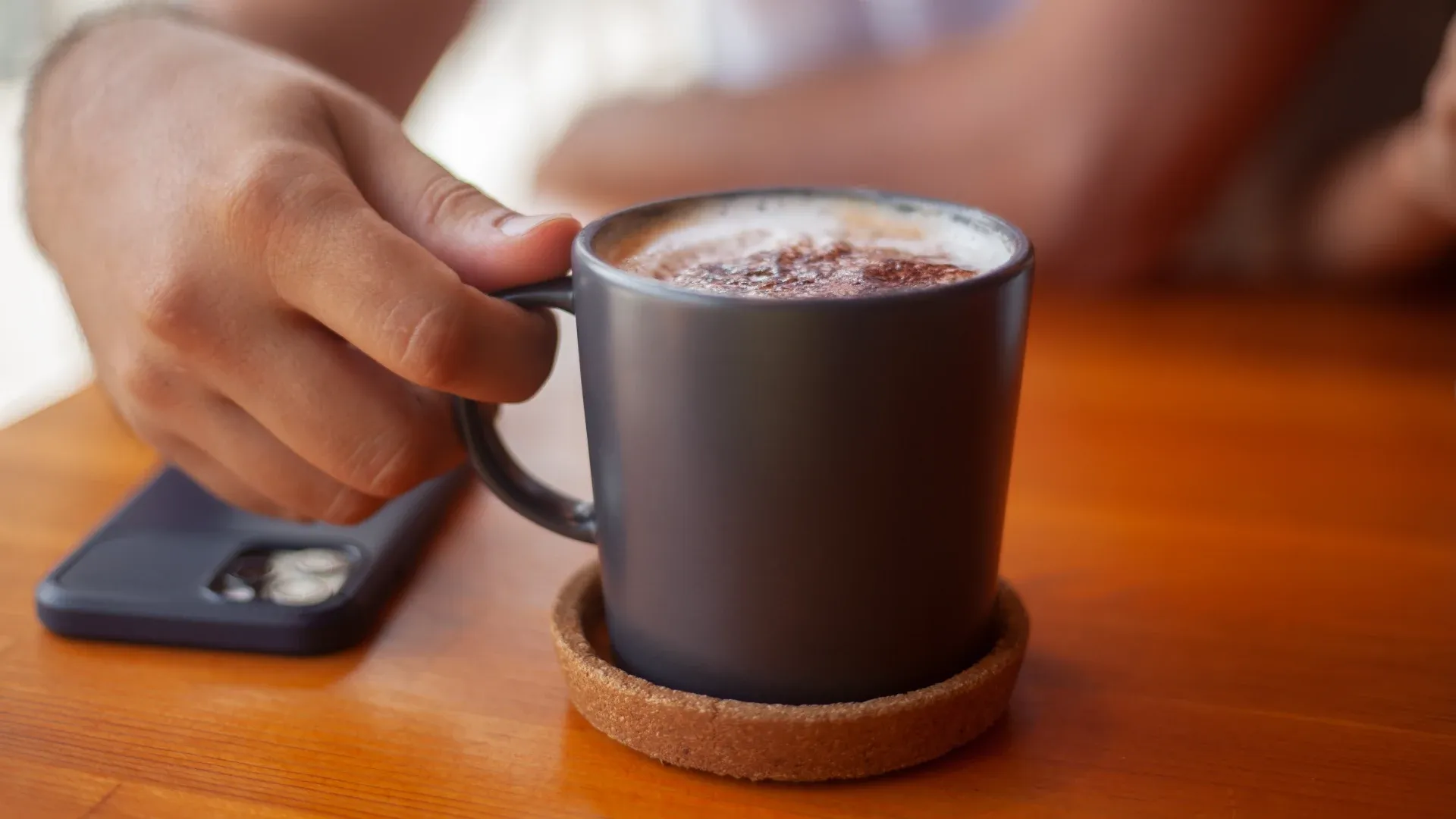 Mão de pessoa negra segurando uma xícara de café apoiada na mesa à luz do dia