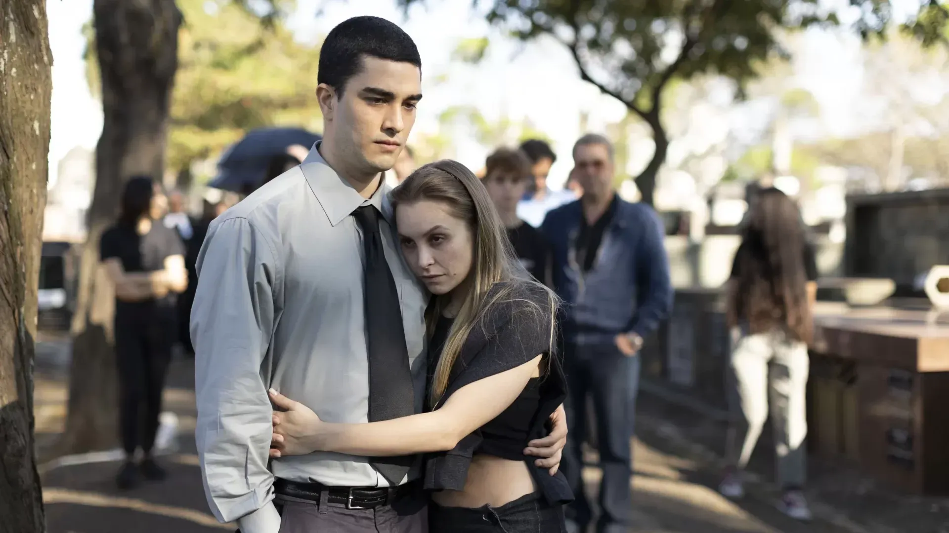 Personagens abraçados com expressões sérias e sem emoção em um velório no fundo