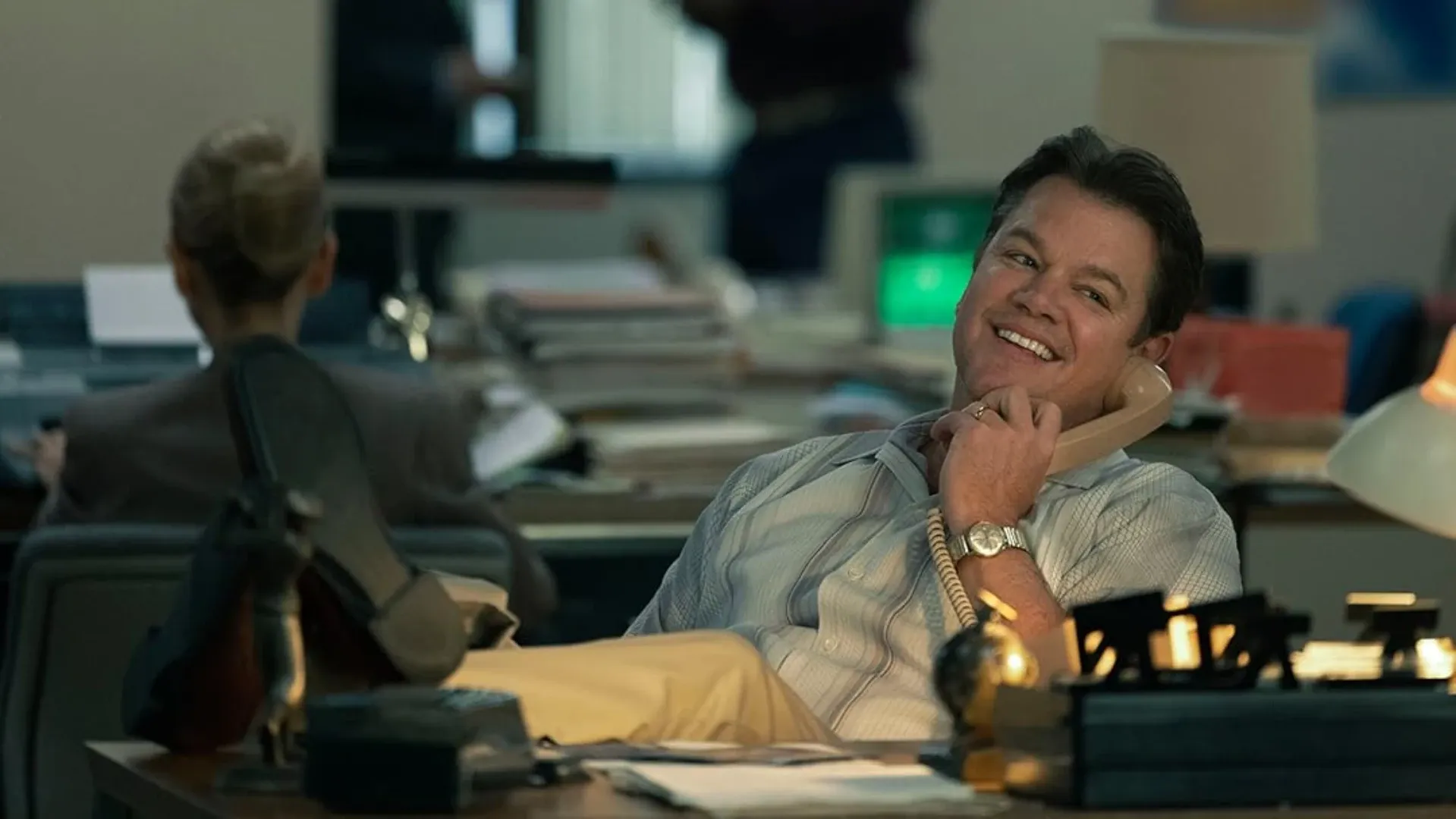 Ator sentado em uma cadeira de escritório com pés na mesa enquanto sorri falando no telefone