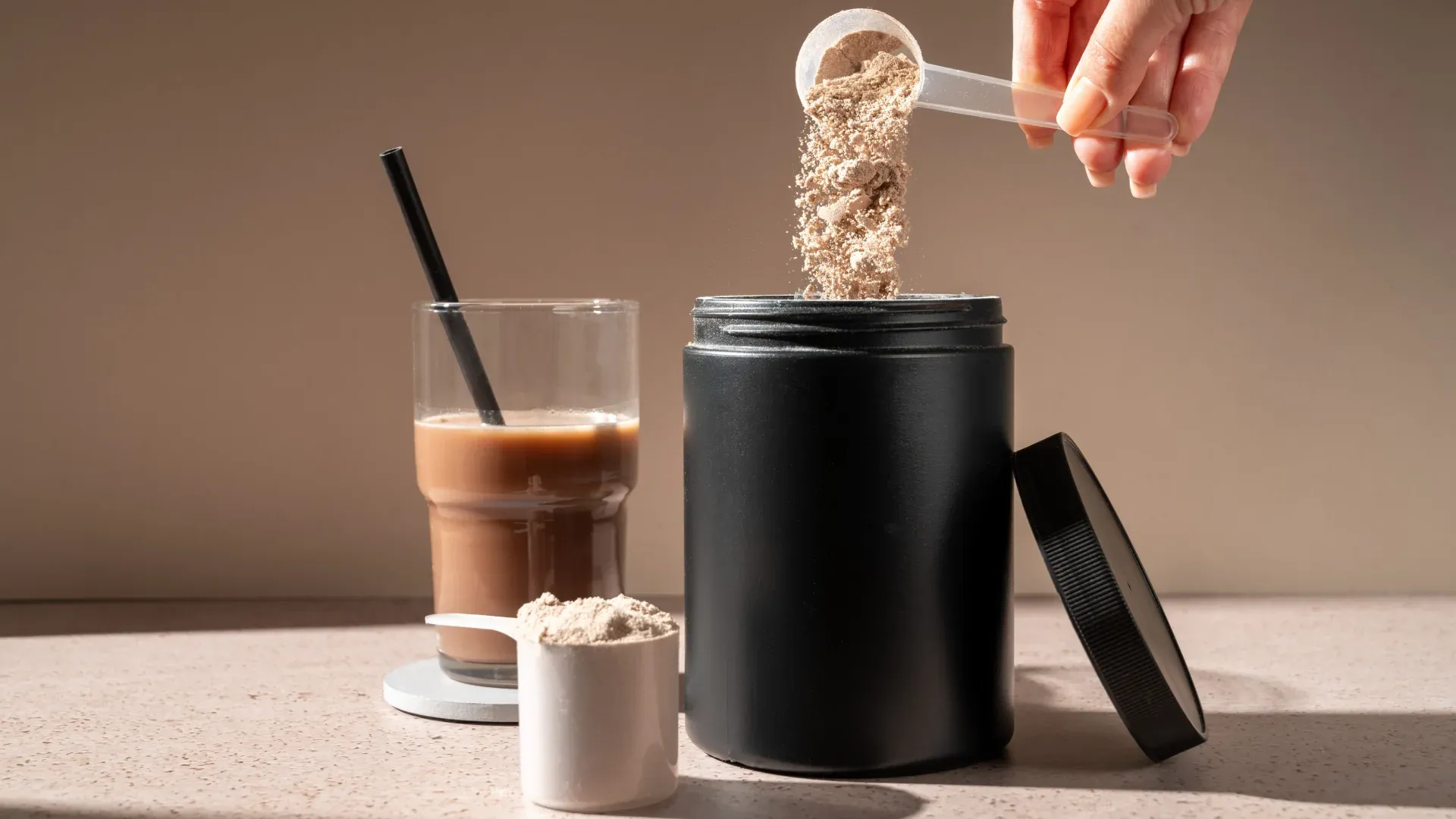 Pote de whey protein com copo de vidro com bebida marrom ao lado e mão segurando scoop