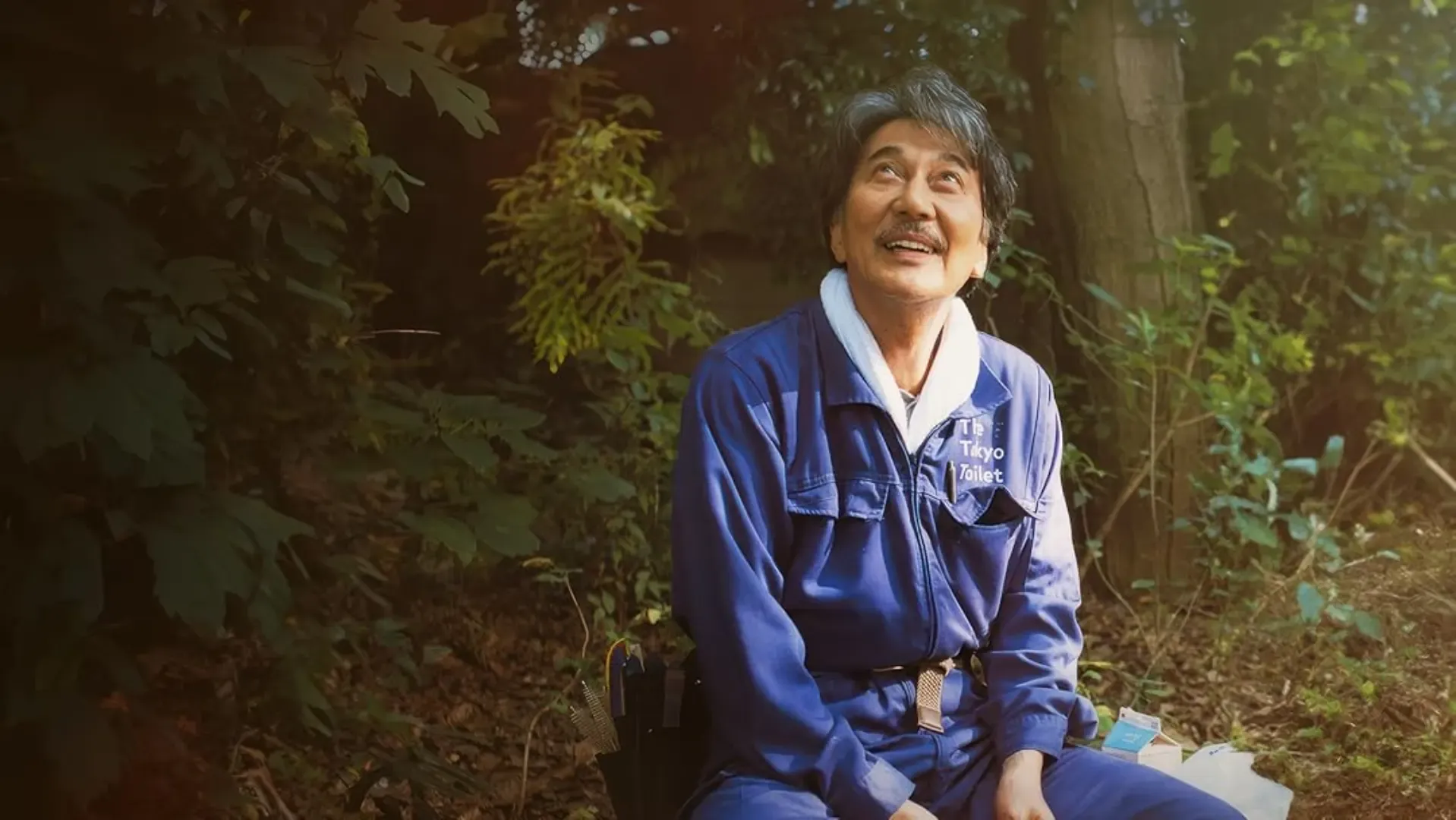 Ator sorrindo enquanto olha para cima em um fundo de paisagem com plantas e árvores