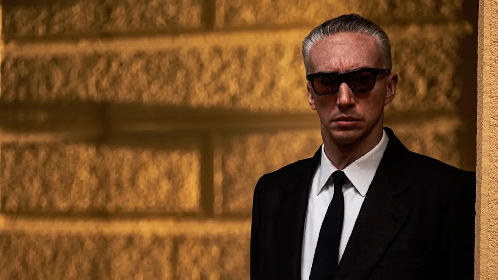 Adam Driver interpretando o personagem com óculos escuros com uma parede laranja meio amarelada no fundo