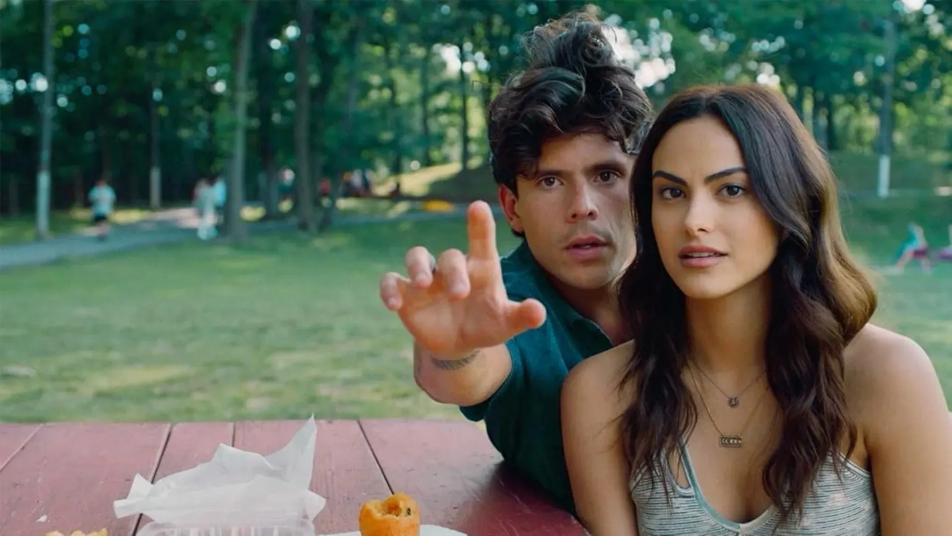 Atores olhando para algo enquanto o homem aponta para alguma direção sentados em um banco num parque