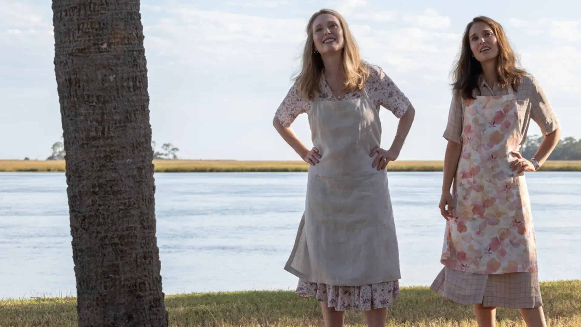 Duas mulheres com aventais olhando para algo ao ar livre com um lago no fundo e um tronco de árvore ao lado