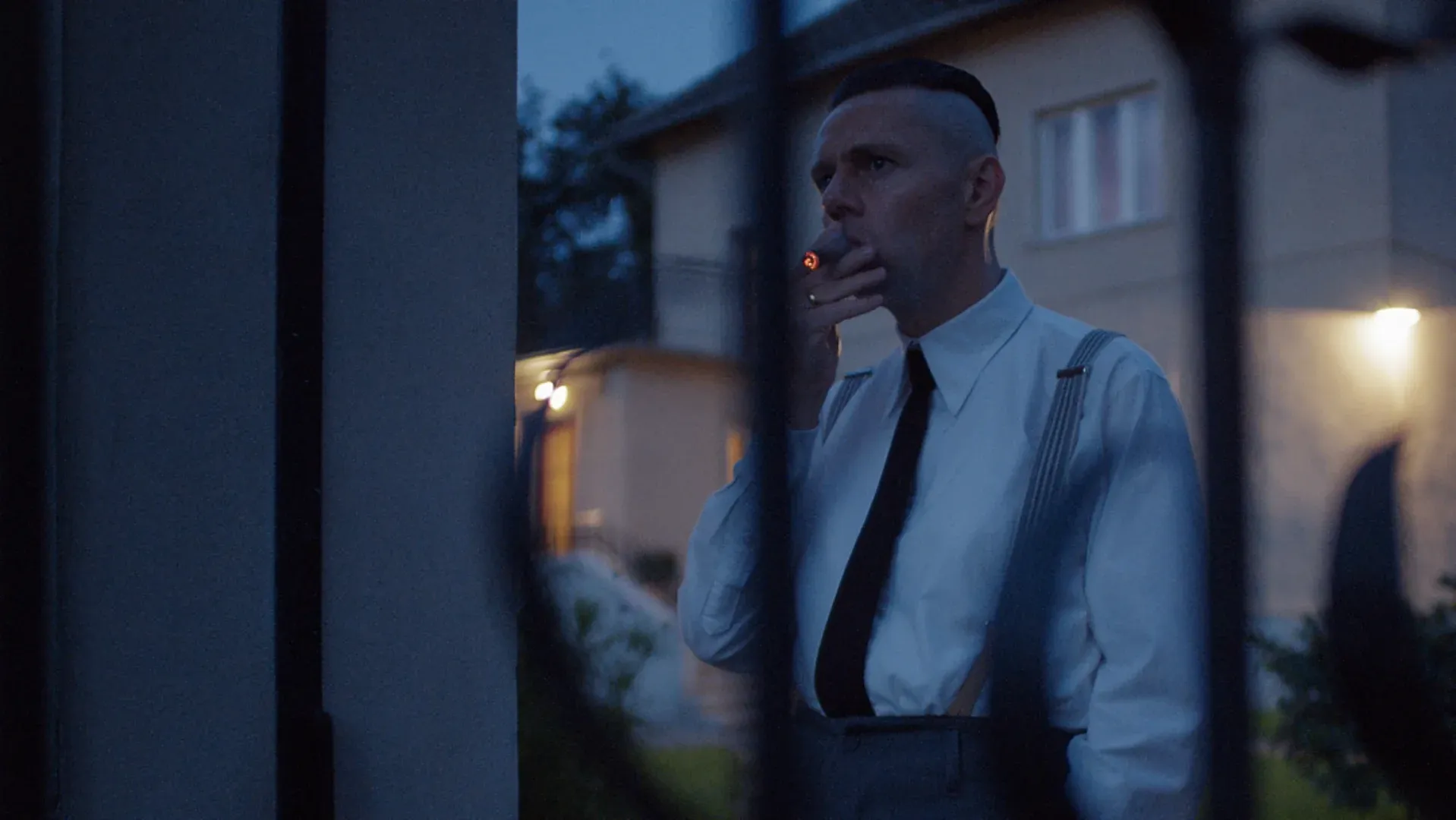 Ator fumando com grades pretas na frente dele com um fundo de uma casa