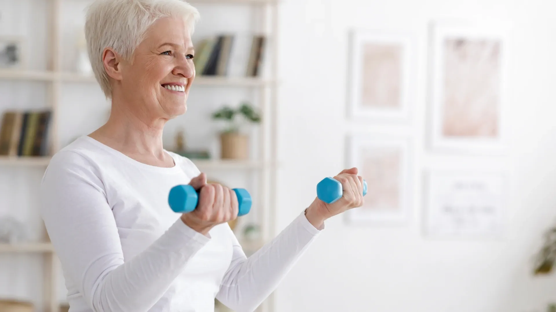 Mulher idosa sorridente realizando exercícios com halteres azuis em casa, vestindo roupas brancas, em um ambiente claro e organizado.
