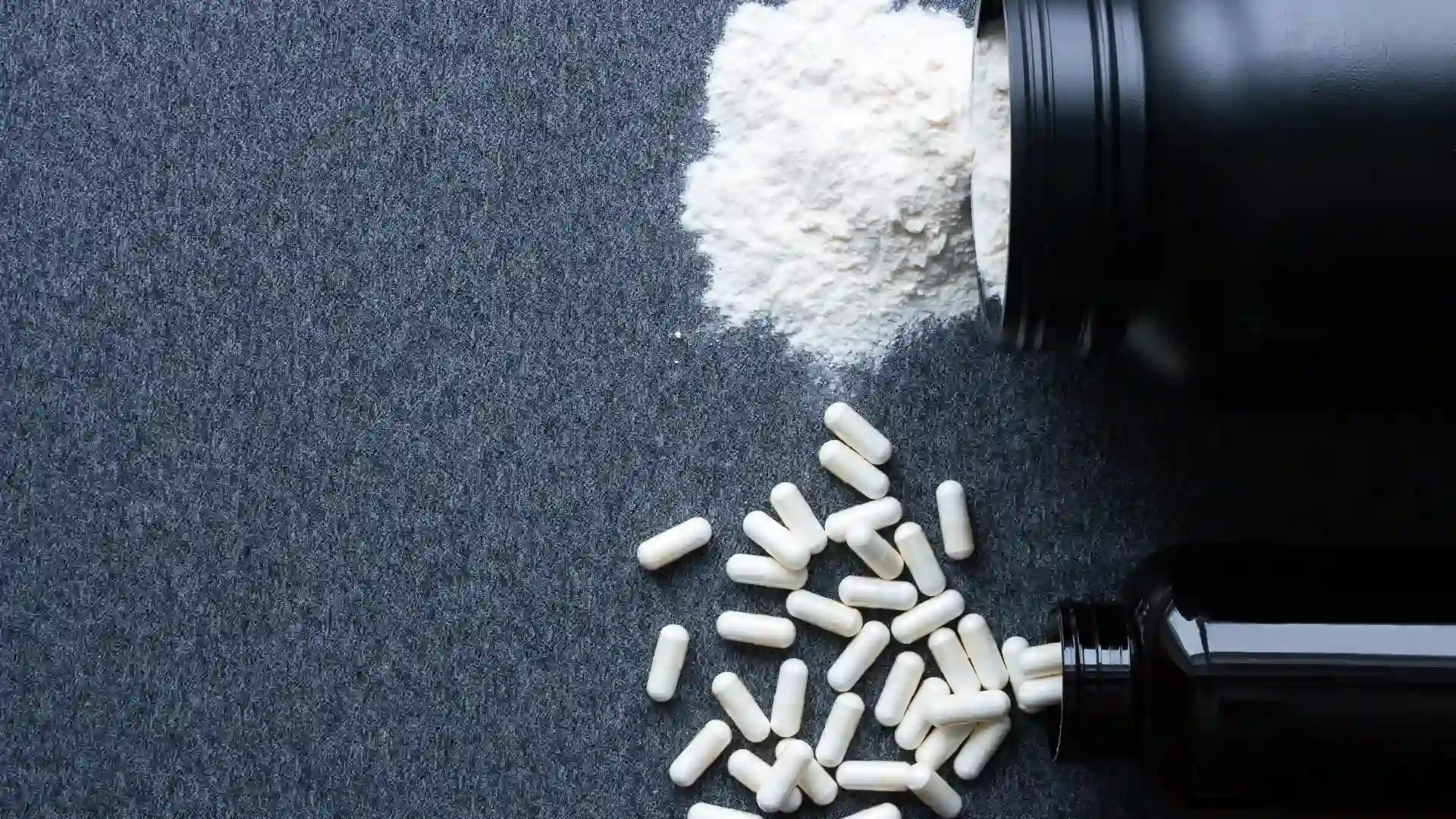 Pote com creatina em pó e um pote com cápsulas de creatina despejadas em uma mesa