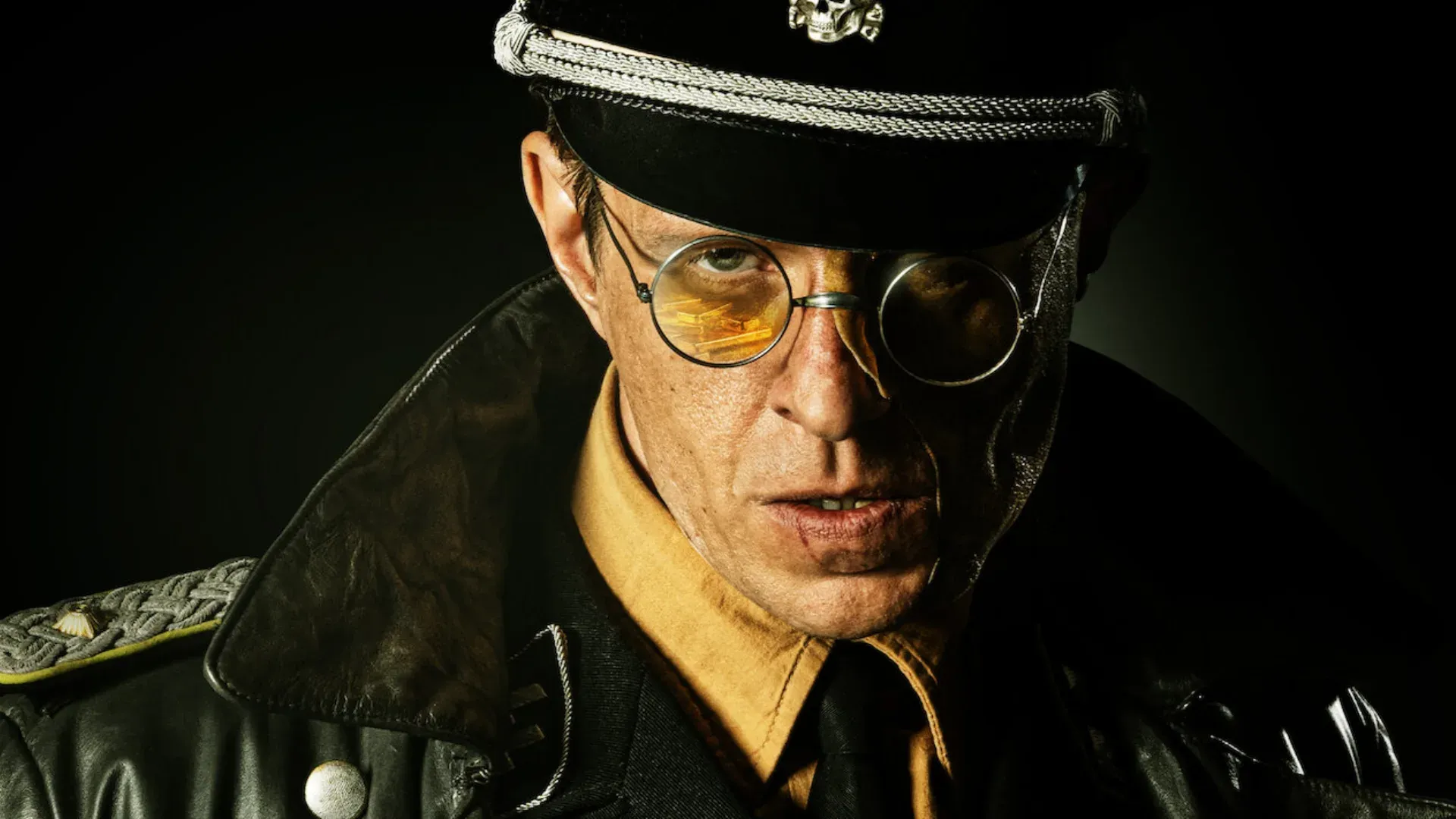 homem com jaqueta de couro, chapéu com uma caveira e um óculos amarelo encarando sério a tela  