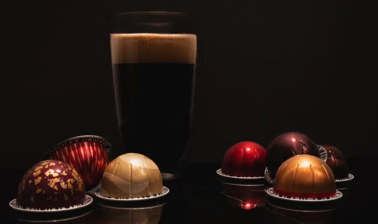 Café preto em uma xícara de vidro com espuma iluminada por cima, e cápsulas coloridas de café Nespresso.