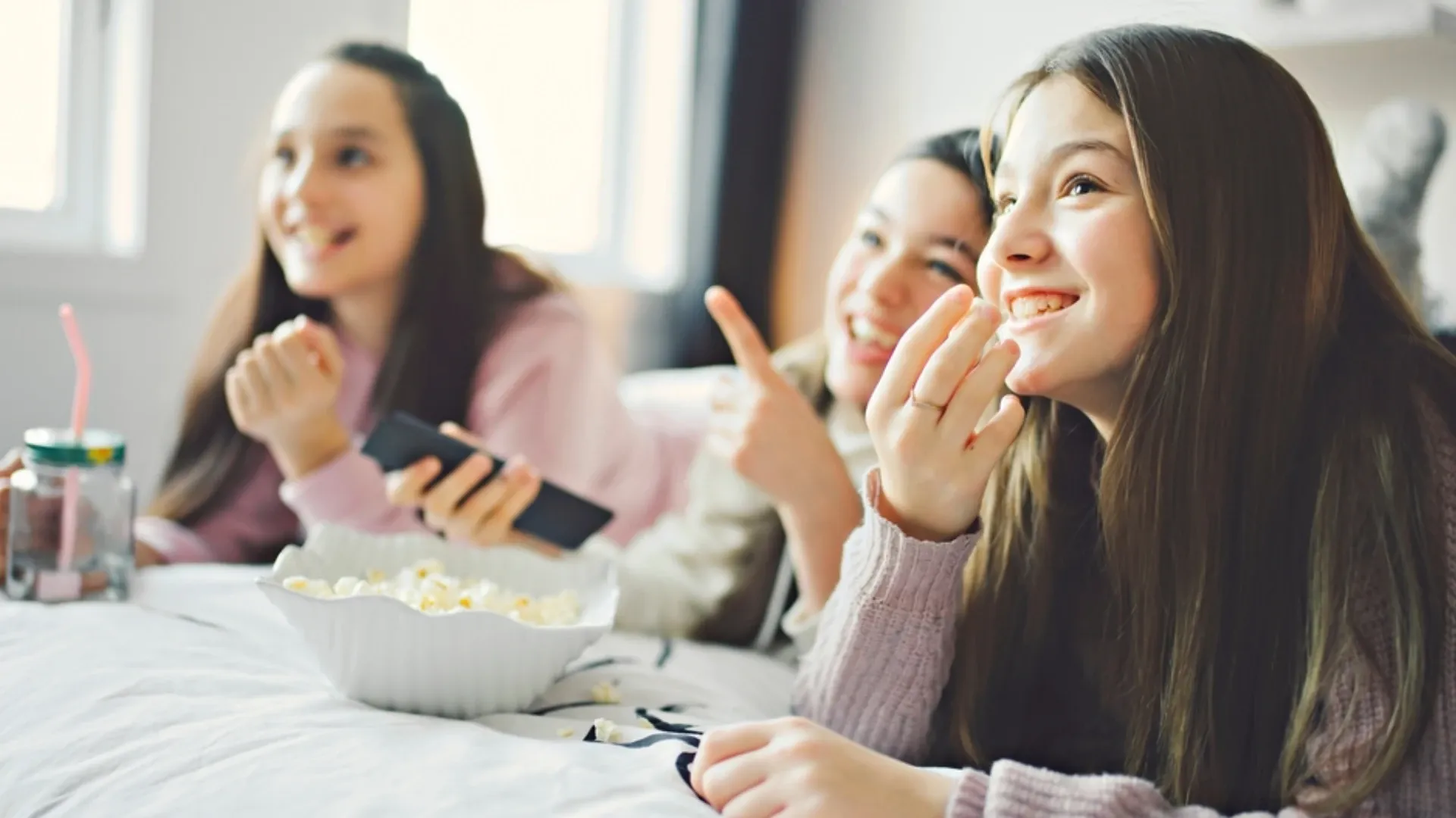 três meninas assistindo TV juntas enquanto sorriem e comem pipoca na cama