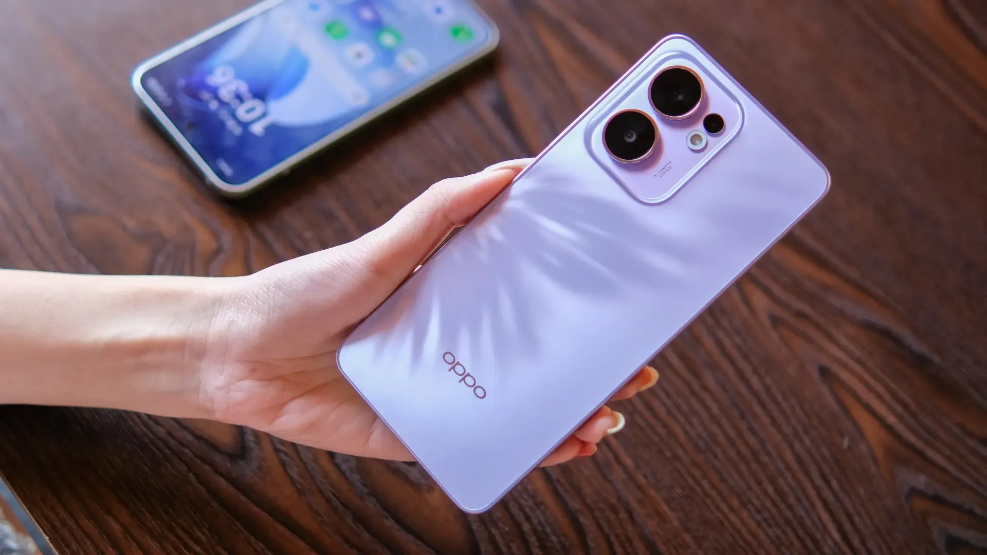 Uma pessoa segura um celular de cor roxa. Ao lado tem uma mesa marrom com outro celular sobre ela