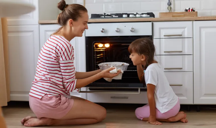 mãe e filha agachadas em frente a fogão de embutir retiram comida de dentro do forno 