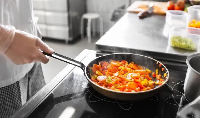 Pessoa cozinhando legumes em uma frigideira no cooktop elétrico.