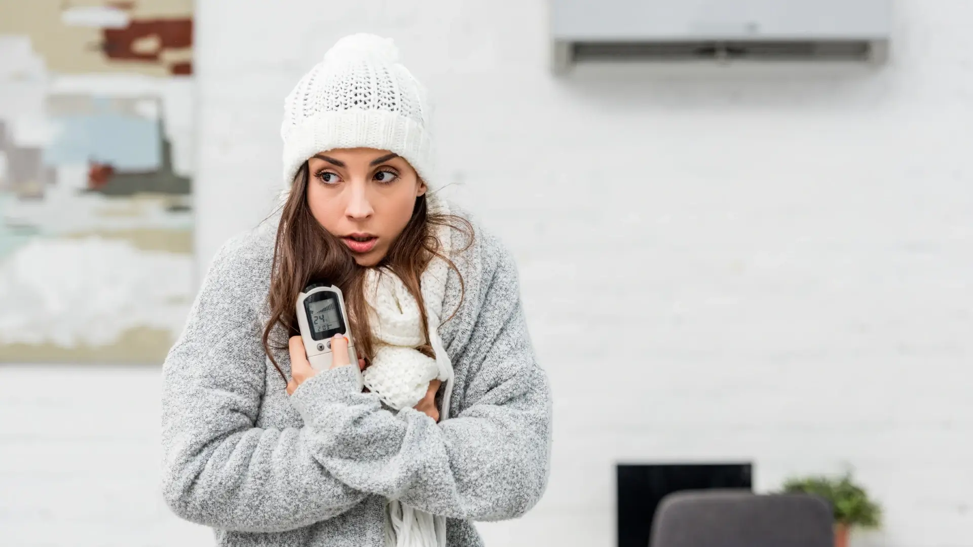 Imagem de uma mulher sentindo frio e segurando o controle de um ar-condicionado que está ao fundo da foto 
